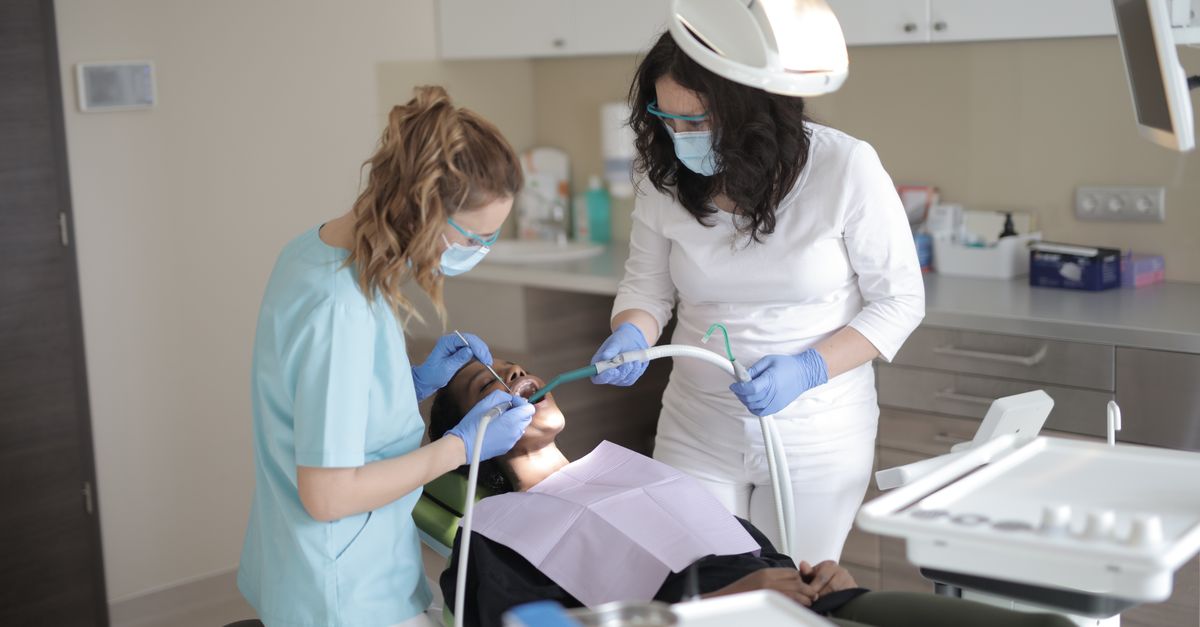 Female dentists in medical masks and latex gloves wearing uniform using medical instruments while treating teeth of female patient in modern dental clinic